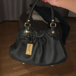 Svart handväska Marc Jacobs - Snygg svart handväska från Marc Jacobs i mjukt skinn med guldfärgade detaljer och metallbricka framtill. Väskan har två handtag, dragkedja och ett coolt mönstrat innerfoder med logotyp. Perfekt storlek för det viktigaste och ger en lyxig vibe. Pris kan absolut diskuteras 