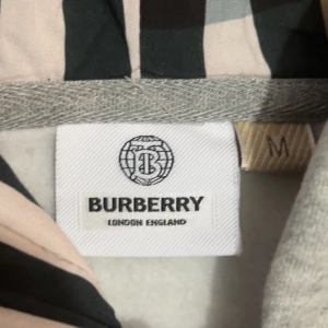 Grå hoodie med dragkedja från Burberry - Snygg ljusgrå hoodie från Burberry med dragkedja framtill, huva med snörning och klassiska fickor. Insidan av huvan har Burberrys ikoniska mönster. Perfekt för en chill och stilren look.