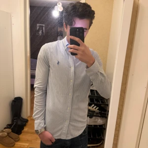 Randig skjorta från Ralph Lauren - Snygg ljusblå och vit randig skjorta från Ralph Lauren med klassisk broderad logga på bröstet. Modellen är slim och har långa ärmar, knappar framtill och krage. Materialet är mjuk bomull.