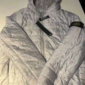 Vit quiltad pufferjacka Stone Island - Säljer en clean vit pufferjacka från Stone Island med huva och dragkedja framtill. Jackan har quiltad design och klassisk patch på ärmen. Materialet är lätt och glansigt, perfekt för dig som vill ha en snygg och modern look. Taggar och etiketter kvar.