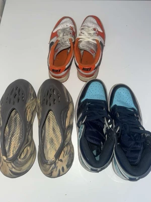 Nike och Yeezy sneakers mix | KOM PM FÖR PRIS! | - | GÅR ATT KÖPA ENSKILDA | KOM PM FÖR INFO + PRIS | Tre par sneakers: ett par Nike Dunk Low i vitt och orange med klassisk swoosh, ett par Adidas Yeezy Foam Runner i grått och beige med futuristisk design och hålmönster, samt ett par Nike Dunk High i svart och ljusblått med vita detaljer och högt skaft.