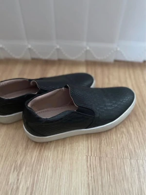 Svarta loafers med ormmönster - Säljer ett par svarta loafers med snyggt ormmönstrat konstläder och vita sulor. Skorna har en stilren slip-on design och är perfekta för dig som vill ha en clean look med lite edge. Passar till det mesta och är superenkla att ta på.