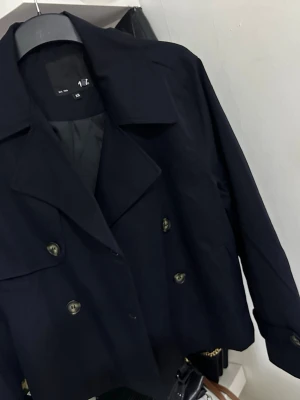 Kort svart trenchcoat från lager 157 - Stilren svart trenchcoat från Lager 157 med dubbelknäppning och klassisk krage. Jackan är kortare i modellen och har snygga knappar framtill samt långa ärmar. Perfekt för dig som gillar minimalistisk och modern stil.