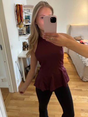 Vinröd peplumtop med hög hals - Säljer en vinröd peplumtop med hög hals och ärmlös design. Toppen har en snygg volang nedtill som ger en cool siluett. Perfekt att matcha med jeans eller kjol för en trendig look.