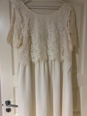 Vit spetsklänning med kort ärm - Romantisk vit klänning med vacker spets upptill och korta ärmar. Klänningen har en lös passform och faller mjukt ner till knäna. Överdelen är dekorerad med blommig spets och underdelen är slät och enkel. Perfekt för sommarens alla tillfällen.