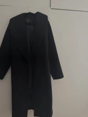 Svart kappa från Vero Moda - Stilren svart kappa från Vero Moda med bred krage och knytskärp i midjan. Kappan har en rak passform och är helfodrad för extra komfort. Perfekt för dig som vill ha en klassisk look med modern touch. Materialet är mjukt och känns lyxigt.