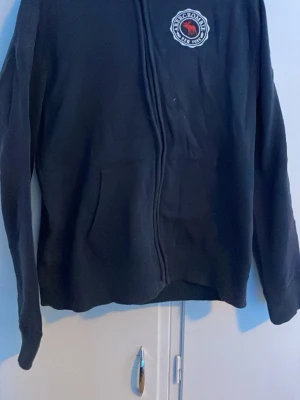 Mörkblå hoodie från Abercrombie - Mörkblå hoodie med dragkedja från Abercrombie. Stor logga med texten 'Abercrombie New York 5th Avenue' och en älg på ryggen, samt mindre logga på bröstet. Klassisk huva och kängurufickor framtill. Perfekt för en avslappnad streetstyle.