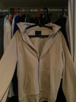 Beige zip hoodie från 157 - Beige hoodie med dragkedja från 157. Klassisk modell med huva och dragsko, samt två fickor framtill. Tillverkad i mjukt och skönt material, perfekt för chill dagar. Enkel och stilren design som passar till det mesta.