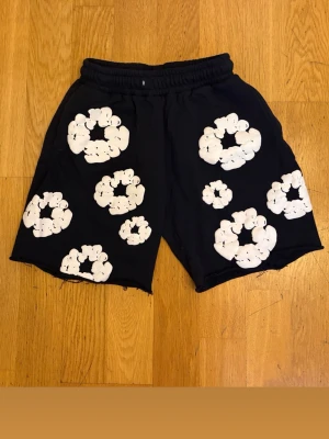 Mörkblåa Denim Tears shorts med vita blommor - Unika mörkblåa Denim Tears med stora vita blommönster över hela plagget. Shortsen har resår i midjan och råa kanter nertill för en avslappnad vibe. Perfekta för sommaren och ger en cool statement-look.