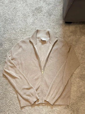 Beige ziptröja från COPE - Beige ribbstickad ziptröja från COPE med hög krage och dragkedja framtill. Tillverkad av återvunnet material, perfekt för lager-på-lager. Tröjan har långa ärmar och ribbade muddar. 