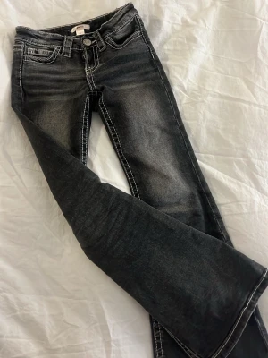 Svarta bootcut jeans med kontrastsömmar - Snygga svarta bootcut jeans med slitningar och vita kontrastsömmar. Jeansen har klassisk femficksdesign och dekorativa sömmar på bakfickorna. Perfekta för dig som gillar en lite retro vibe och vill sticka ut med detaljer.