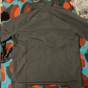 Mörkgrå Nike vindjacka med huva - Snygg mörkgrå vindjacka från Nike med hel dragkedja framtill, huva och två sidofickor. Jackan har en minimalistisk design med Nike-logga på bröstet och reflekterande detaljer på ärmarna. Perfekt för blåsiga dagar och sportiga outfits.