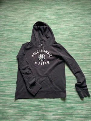 Mörkgrå hoodie Abercrombie & Fitch - Mörkgrå hoodie från Abercrombie & Fitch med broderad logga och text på bröstet. Klassisk känguruficka framtill och justerbar dragsko i huvan. Tillverkad i mjuk bomullsblandning som känns skön mot huden. Perfekt för en avslappnad och cool stil.passar dig runt 175 cm. Står storlek m i men den passar som S