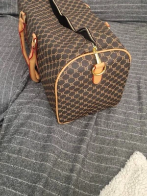 Väska  - Snygg brun väska i klassiskt Triomphe-mönster i beige. Väskan har ljusbruna läderhandtag och detaljer, samt dragkedja i guld. Perfekt storlek för resor och har en stilren, boxig form. Materialet är tåligt canvas med läderdetaljer. Använd 1 gång 