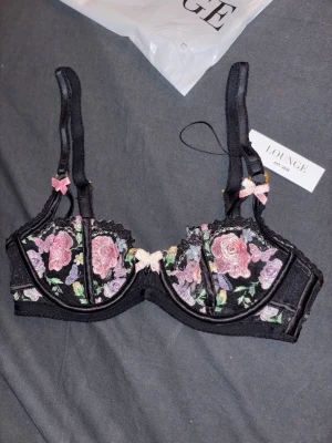 Svart spets-bh med blommor från Lounge - Säljer en svart bh från Lounge med vackra broderade blommor i rosa, lila och grönt. Bh:n har spetsdetaljer, små rosa rosetter och justerbara axelband. Det är storlek 65b och prislappen sitter på. 