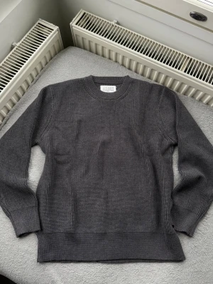Grå stickad tröja Maison Margiela - Säljer en stilren grå stickad tröja från Maison Margiela. Tröjan har rund halsringning, ribbade muddar och är tillverkad i ett tjockare, mjukt material som passar perfekt för kyligare dagar. Enkel och tidlös design som är lätt att matcha.