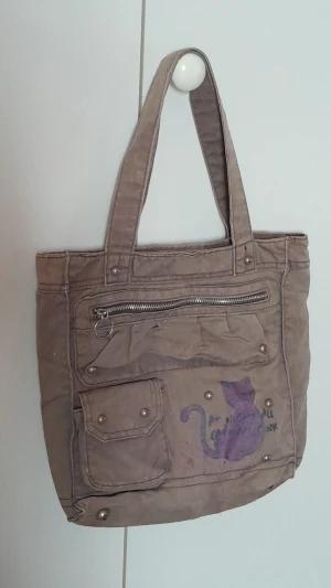 Brun väska - En brun väska med flera fack, dragkedjor och tryck på framsidan.