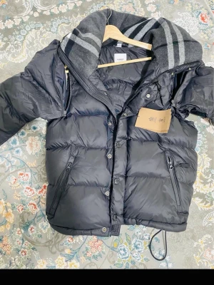 Svart dunväst från MONCLER - Snygg svart dunväst från MONCLER med stor, vadderad krage och gråa ränder. Västen har två dragkedjefickor fram och stängs med både dragkedja och knappar. Perfekt för lager-på-lager och riktigt trendig look.