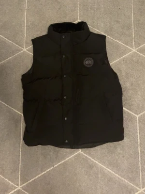 Svart dunväst från Canada Goose - Snygg svart dunväst från Canada Goose med hög krage och tryckknappar framtill. Västen har två sidofickor och en diskret logotyp på bröstet. Perfekt för lager-på-lager och håller dig varm under kyliga dagar.