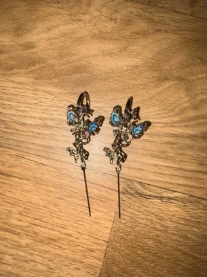 Silvriga örhängen med blå detaljer - Unika örhängen i silverfärgad metall med fantasifullt formade detaljer och inslag av blå emalj. Designen har en konstnärlig och lekfull känsla med långa, smala hängen. Perfekt för dig som vill sticka ut och addera färg till din stil.