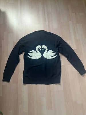 Mörkblå stickad tröja med svanar - Mysig mörkblå stickad tröja med två vita svanar som bildar ett hjärta på bröstet. Tröjan har rund halsringning och långa ärmar. Stor i storleken men väldigt fin.