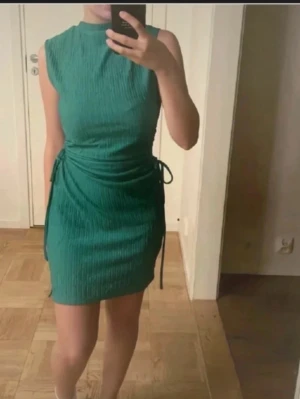 Grön ribbad kort klänning - Snygg grön kortklänning med ribbad struktur och snörning i sidan. Ärmlös modell med hög hals och figurnära passform. Perfekt för dig som vill ha en enkel men trendig look.