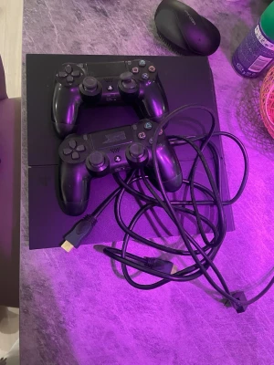 PlayStation 4 med två handkontroller - Svart PlayStation 4 med två svarta DualShock 4-handkontroller och tillhörande kablar. Konsolen har en stilren, rektangulär design i matt plast och är perfekt för gamingkvällar med vänner. HDMI-kabel och strömkabel ingår.