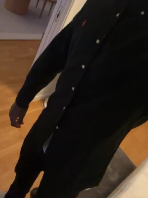 Svart skjorta från Ralph Lauren - Snygg svart skjorta från Ralph Lauren med klassisk krage och knäppning framtill. Skjortan har en röd broderad logga på bröstet och vita knappar. Perfekt för en stilren look och passar till många olika outfits.