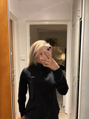 Svart sporttröja med dragkedja - Svart sporttröja med halv dragkedja framtill och liten logga på bröstet. Tröjan har lång ärm, hög krage och är tillverkad i ett mjukt, stretchigt material som passar perfekt till både träning eller vardag. 