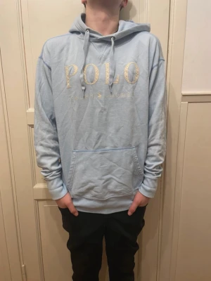 Ljusblå hoodie från Polo Ralph Lauren - Ljusblå hoodie från Polo Ralph Lauren med stor gul logotyp på bröstet. Hoodien har huva med dragsko och en stor magficka. Mjuk och skön bomullsmix som passar perfekt till chill dagar. Klassisk och stilren design som är enkel att matcha.