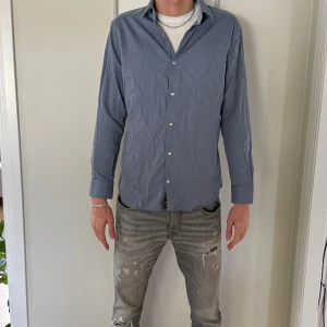 Blå slim fit skjorta från Riley - Snygg blå skjorta från Riley i slim fit-modell. Skjortan har diskreta vita ränder, klassisk krage och knäppning framtill. Långärmad och tillverkad i mjuk bomull, perfekt för en clean och stilren look.