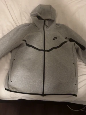 Grå Nike Tech Fleece hoodie - Snygg grå Nike Tech Fleece hoodie med svart dragkedja och svarta detaljer. Hoodien har huva, två sidofickor med dragkedja och en modern, sportig look. Perfekt för dig som gillar streetwear och vill ha något bekvämt och stilrent.