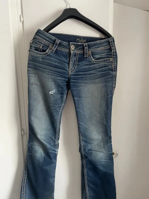 Blå bootcut jeans från Silver Jeans - Snygga blå bootcut jeans från Silver Jeans med slitningar och ljusa partier. Jeansen har dekorativa bakfickor med kontrastsömmar och klassisk femficksdesign. Köparen står för frakten 