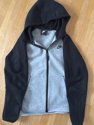 Nike Tech old - Nike hoodie med dragkedja i grått och svarta ärmar samt huva. Klassisk logga på bröstet och två fickor framtill. Perfekt för en sportig och avslappnad stil. Materialet är mjukt och bekvämt, passar till både träning och chill.