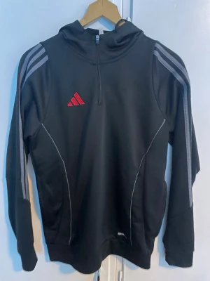 Svart Adidas hoodie med half zip - Svart hoodie från Adidas med grå ränder på axlarna och rött Adidas-logo på bröstet. Tröjan har en praktisk half zip och huva, samt vita detaljer längs sidorna. Perfekt för dig som gillar sportig stil.