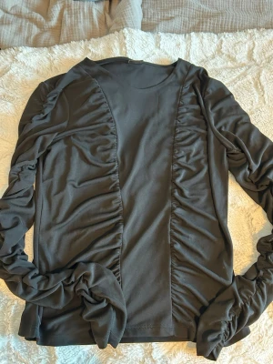 Svart långärmad topp från Gina Tricot - Snygg svart långärmad topp från Gina Tricot med rynkade detaljer längs sidorna och ärmarna. Tillverkad i mjukt och stretchigt material som sitter bekvämt. Perfekt att styla med jeans eller kjol för en trendig look.