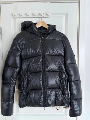 Save The Duck Edward - Säljer en svart pufferjacka från Save The Duck med huva och dragkedja framtill. Jackan är tillverkad i glansigt, djurfritt material och har två fickor med dragkedja. Perfekt för kalla vinterdagar och riktigt lätt att bära.