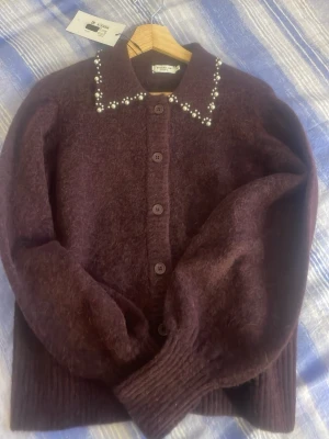 Vinröd kofta med pärldetaljer - Mysig vinröd kofta från SisterS Point med stora knappar framtill och dekorativa pärlor längs kragen. Tillverkad i fluffigt material som ger en lyxig känsla. Perfekt för dig som vill ha något extra till din outfit.