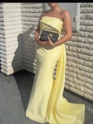 Lång gul axelbandslös klänning - Elegant axelbandslös långklänning i ljusgul färg med släp. Klänningen har en slimmad passform och är tillverkad i ett glansigt material som ger en lyxig känsla. Perfekt för speciella tillfällen och högtider.