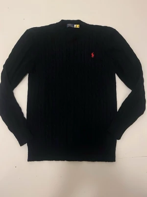 Svart kabelstickad tröja Polo Ralph Lauren - Kabelstickad svart tröja från Polo Ralph Lauren med rund halsringning och den klassiska röda logotypen broderad på bröstet. Tröjan har långa ärmar och ribbade muddar vid ärmslut och nederkant. Perfekt för en stilren och tidlös look.