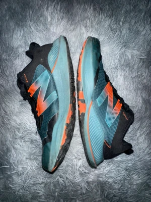 New Balance blå och orange sneakers - Snygga sneakers från New Balance i blått och svart med stora orange detaljer på sidorna. Skorna har en sportig design med mesh och syntetmaterial, samt robust sula med orange inslag. Perfekta för dig som gillar streetwear och vill sticka ut.