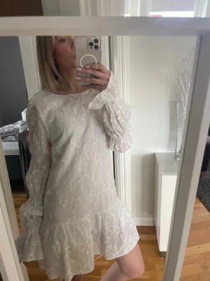 Vit blommig kortklänning med volang - Supersöt vit kortklänning med blommigt broderat mönster och volang nertill. Klänningen har långa, lätt utsvängda ärmar och rund halsringning. Perfekt för sommarens alla tillfällen och ger en romantisk vibe.