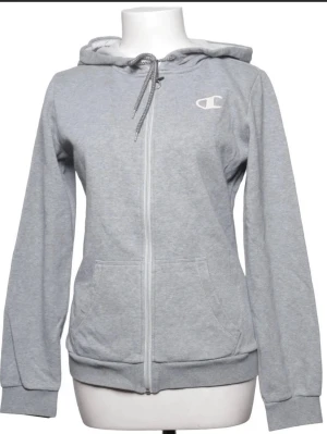 Grå zip-hoodie från Champion - Snygg grå zip-hoodie från Champion med huva och dragsnören. Klassisk logga på bröstet, två fickor framtill och ribbade muddar vid ärmslut och nederkant. Perfekt för en chill look och enkel att matcha med jeans eller joggers.
