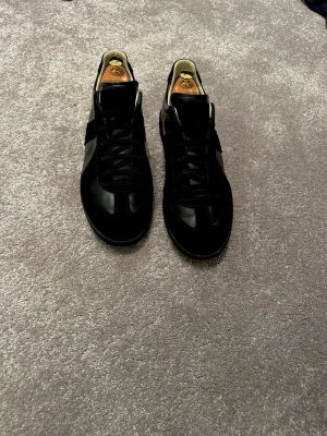 Maison Margiela gats  - Säljer ett par stilrena svarta maison margiela i mocka med klassisk låg profil och svarta skosnören. Insidan är ljus och skon har en enkel design som passar till många olika outfits. Perfekt för dig som gillar stilrena outfits. Vid frågor eller funderingar, tveka inte på att skriva! 