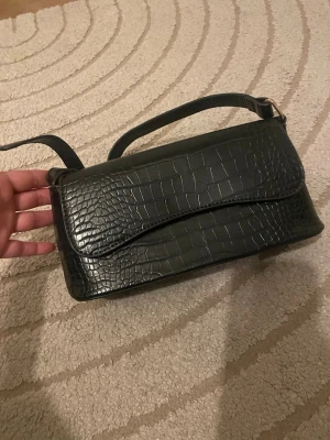 Svart axelväska i krokopräglat material - Snygg svart axelväska med krokodilpräglat mönster och justerbar axelrem. Väskan har en rektangulär form och stängs med lock. Perfekt för dig som gillar trendiga accessoarer med lite edge.