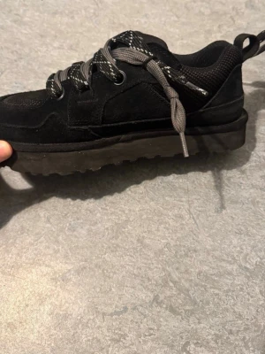 Svarta sneakers från UGG - Snygga svarta sneakers från UGG med robust sula och gråsvarta mönstrade skosnören. Ovandelen är i mocka och mesh för en cool look och bra andningsförmåga. Perfekta för dig som gillar streetstyle och vill ha något bekvämt och stilrent.