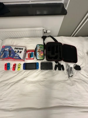 Nintendo Switch Fortnite Edition med tillbehör - Nintendo Switch Fortnite Edition med blå och röda Joy-Con, extra neon Joy-Con, laddare, dockningsstation, handkontrollhållare, Fortnite-låda, Mario-väska och svart bärväska. Perfekt för gaming hemma eller på språng.