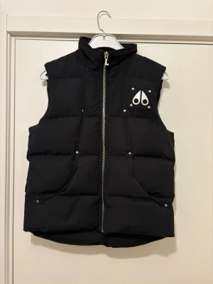 Svart dunväst från Moose Knuckles - Snygg svart dunväst från Moose Knuckles med hög krage och silverfärgad dragkedja. Västen har två framfickor med tryckknappar och en ikonisk logga på bröstet. Perfekt för lager-på-lager och håller dig varm under kyliga dagar.