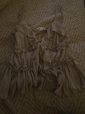 Beige volangtop med smala axelband - Supersöt beige linnetopp med volanger och smala axelband. Toppen har plisserat tyg, knytning framtill och flera lager volangdetaljer som ger en flowy känsla. Nypris 200kr💕💕