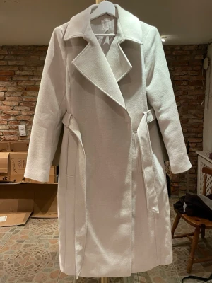 Cremefärgad lång kappa med skärp - Cremevit/beige kappa med bred krage och knytskärp i midjan. Kappan har en rak passform, långa ärmar och två sidofickor. Funkar till allt och knappast använd 🌻